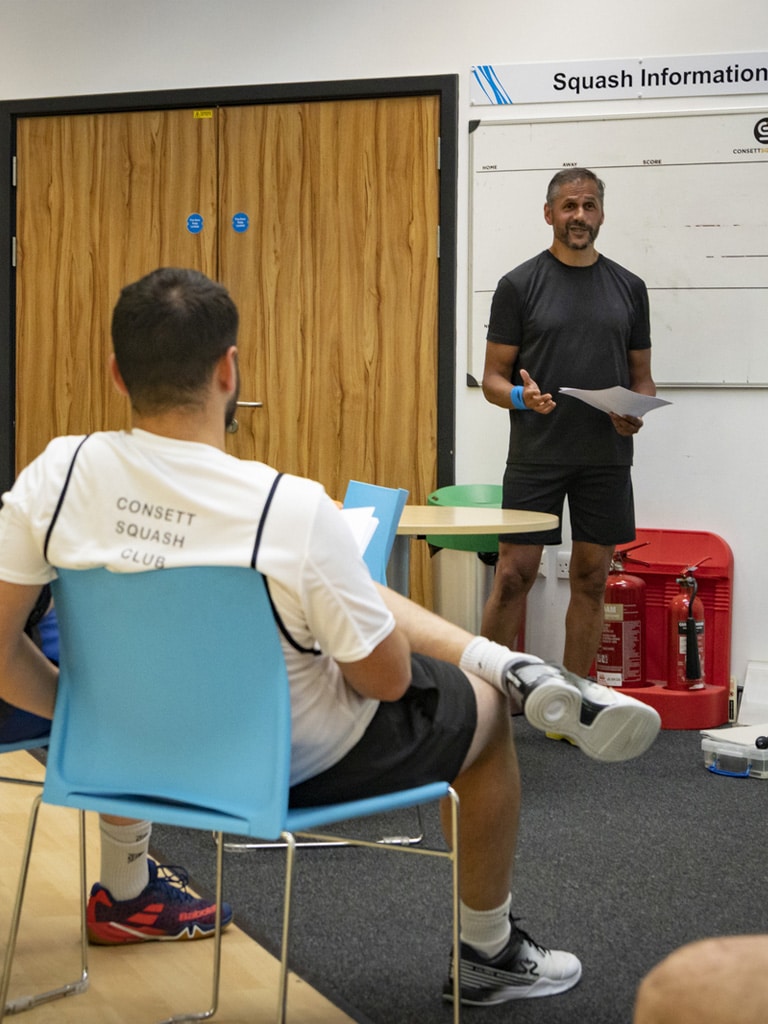 Consett Squash Club written on the back of a man's shirt whilst they listen to a presentation.